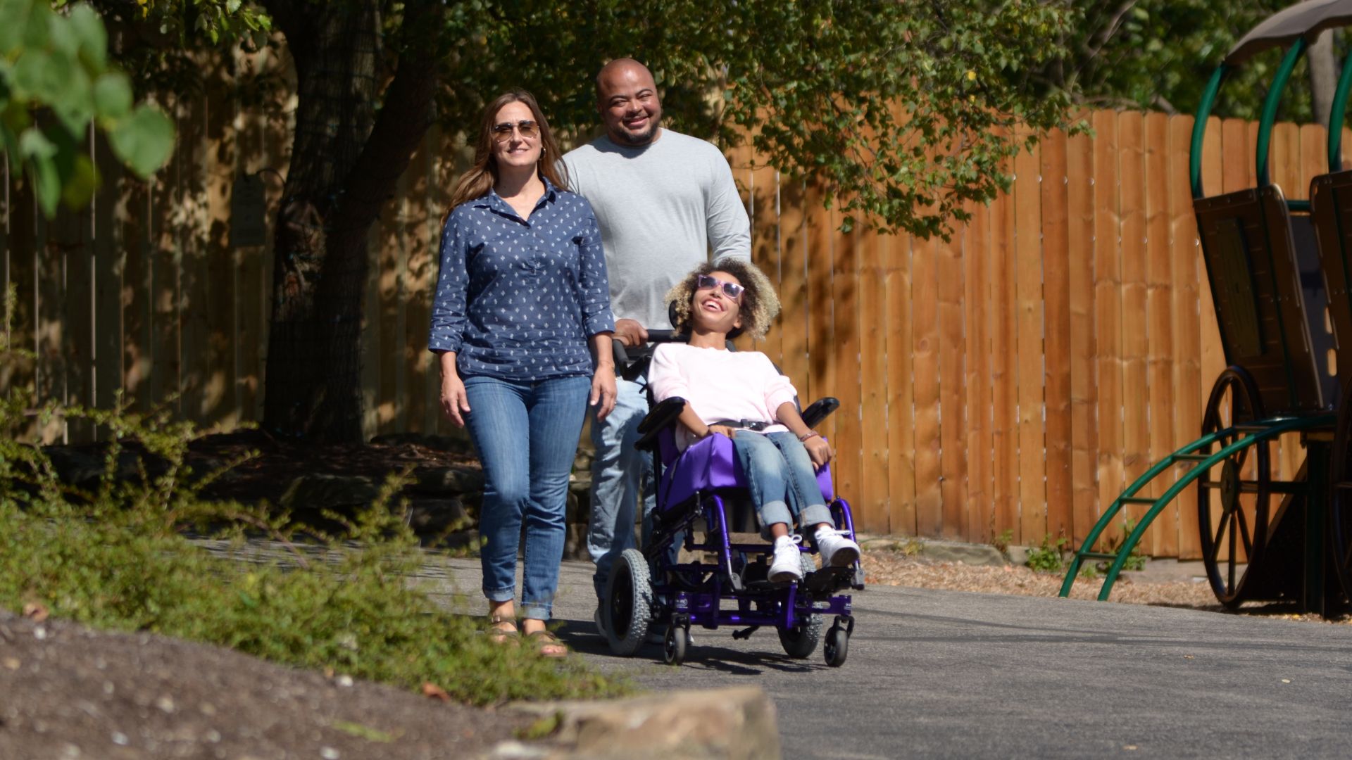 A smiling family walks outdoors on a sunny day; a woman walks beside a man who is pushing a young girl in a wheelchair with advanced Complex Rehab Technology along a paved path, with trees and a wooden fence in the background.