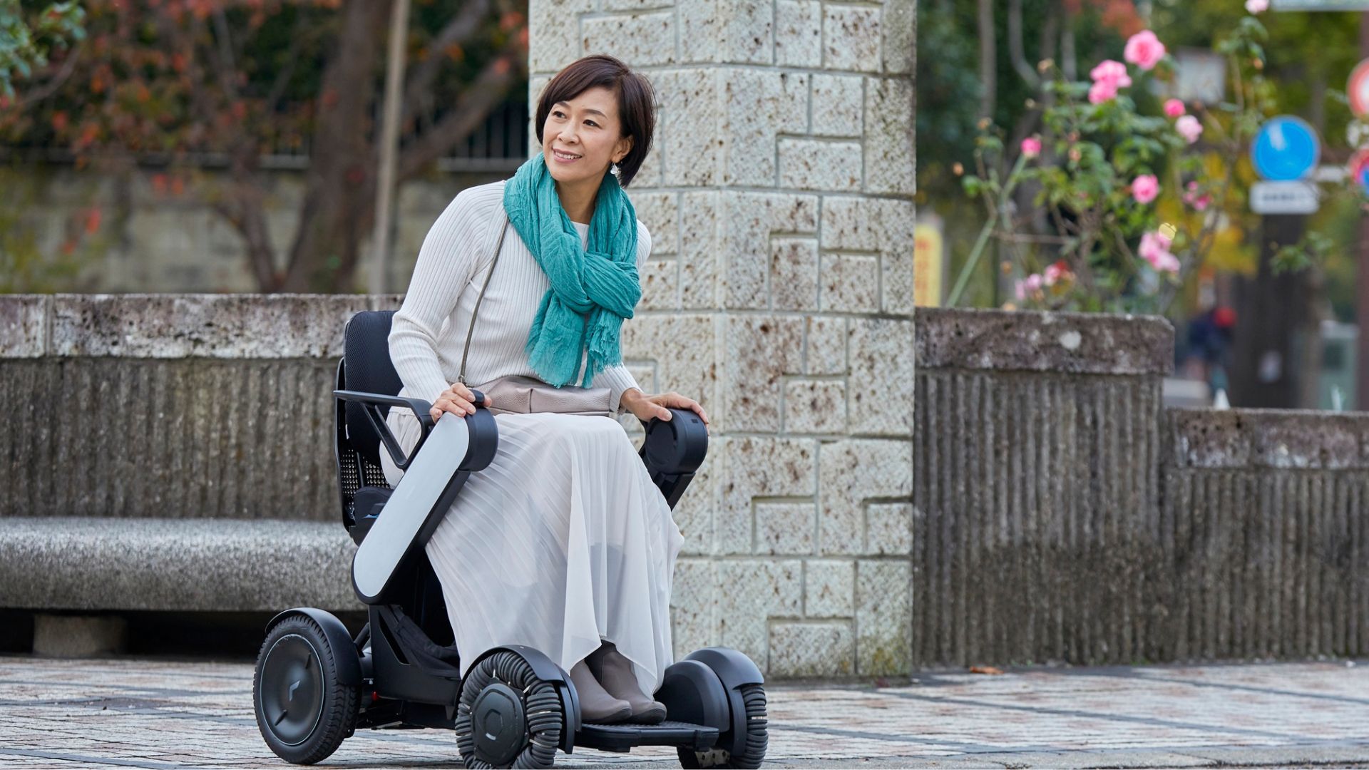 A woman in a white dress and teal scarf smiles while sitting in a modern electric wheelchair system outdoors, surrounded by stone structures and blooming flowers.