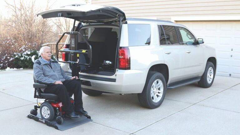 An older man in a motorized wheelchair sits on a wheelchair lift with tilt recline next to an SUV with its rear hatch open, showing the powered seating and lift mechanism for loading the chair into the vehicle.