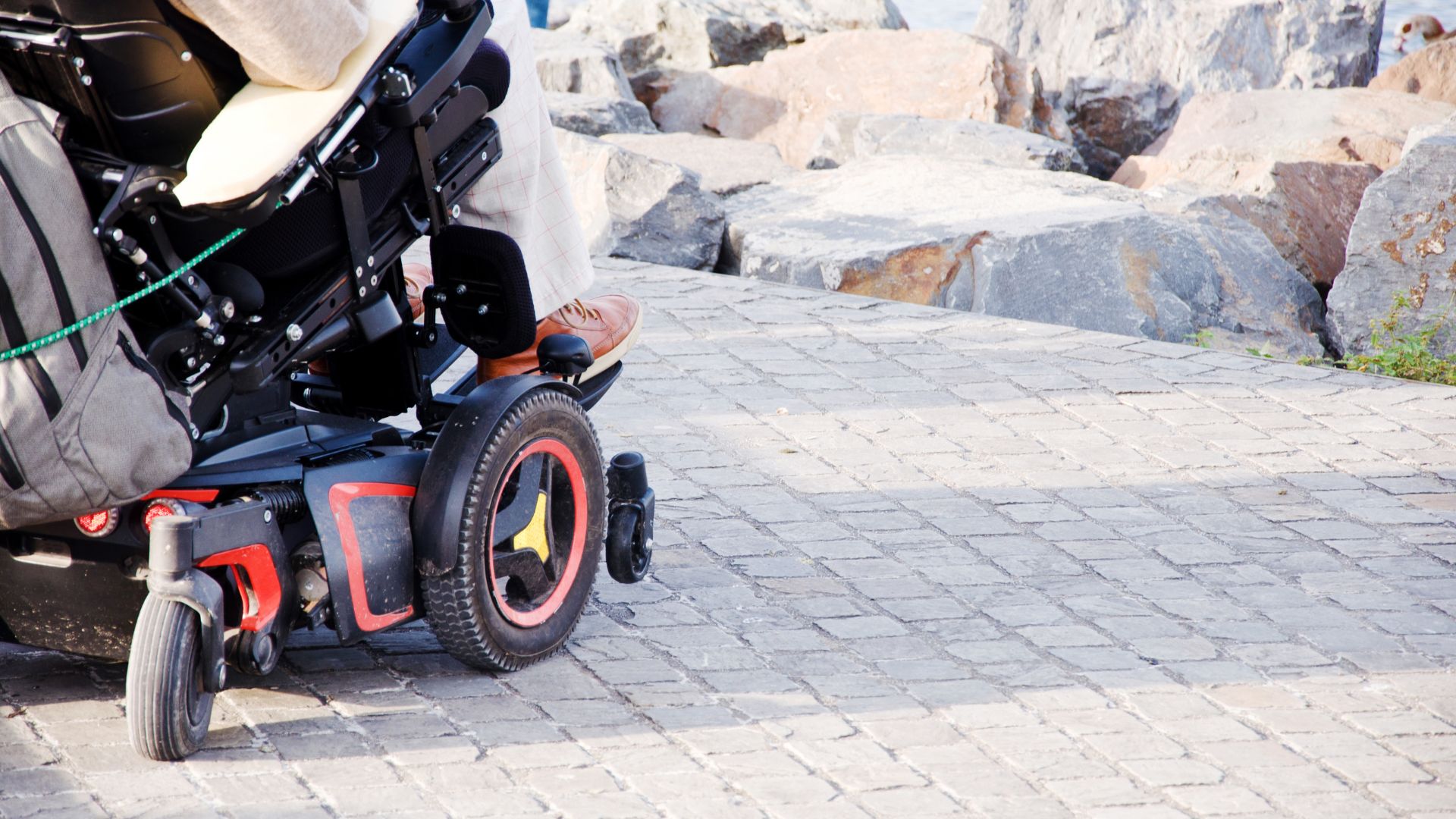 A person in a powered wheelchair, utilizing complex rehab technology, moves along a paved pathway near large rocks, with only the lower part of the wheelchair and the person’s legs visible.