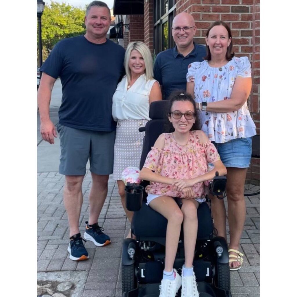 Five adults, one seated in a motorized wheelchair, pose and smile together outdoors on a brick sidewalk near a building. The group appears happy, sharing camaraderie as they celebrate Kierras Story together.