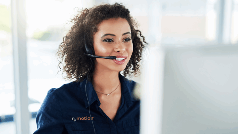 A woman with curly hair wearing a headset and a navy Nu Motion shirt is smiling while looking at a computer screen in a bright, modern office, assisting Medicare beneficiaries with wheelchair prescribing.