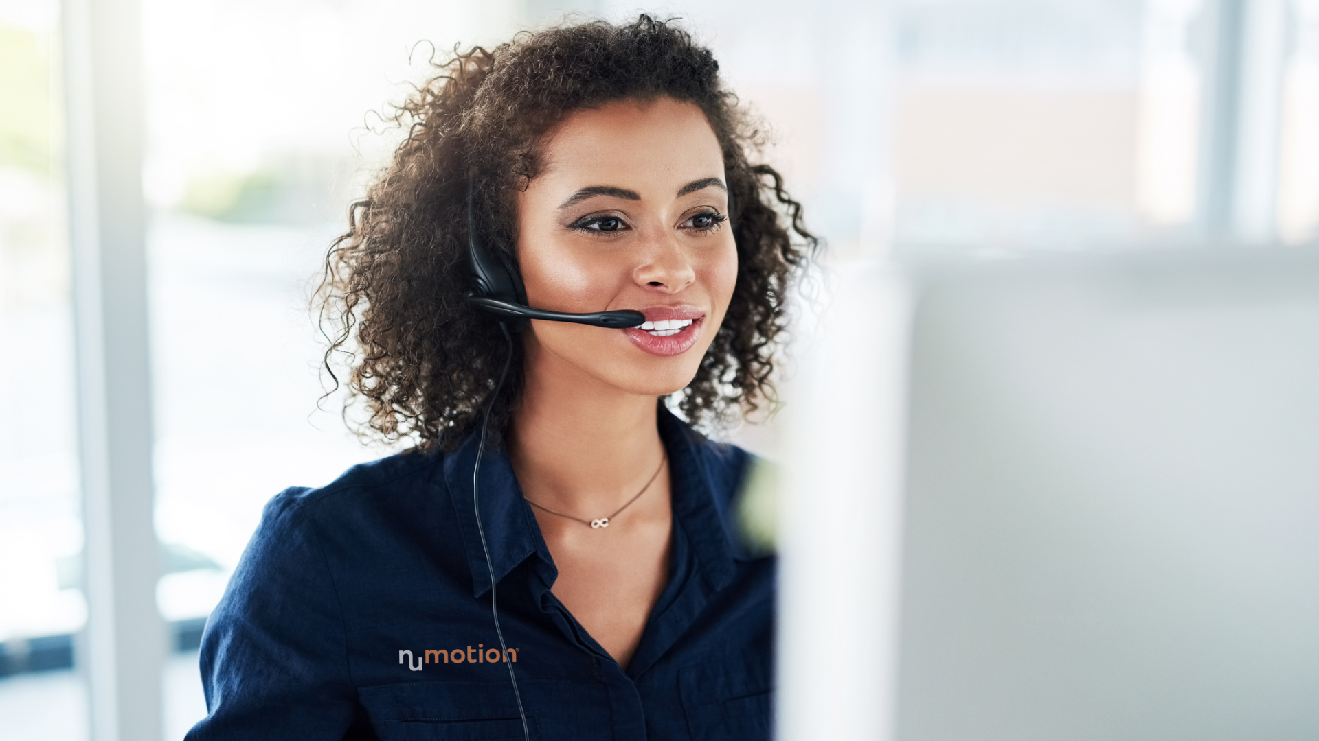 A woman with curly hair wearing a headset and a navy Nu Motion shirt is smiling while looking at a computer screen in a bright, modern office, assisting Medicare beneficiaries with wheelchair prescribing.