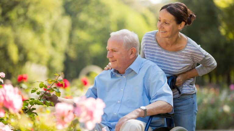 An elderly man aging with disabilities enjoys a garden as a smiling woman stands behind him, gently holding the wheelchair. They are surrounded by blooming flowers and greenery on a sunny day.