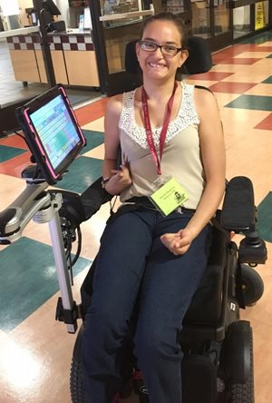 A woman smiling in a powered wheelchair with a mounted tablet device shares Arcys Story. She wears glasses, a sleeveless top, jeans, and a lanyard with a name badge. The background shows a tiled floor and building interior.