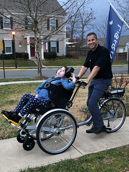 A man smiles while pushing a child in a wheelchair-adapted tricycle on a sidewalk in a suburban neighborhood. The child, featured in Carolines Story, wears a blue outfit with yellow spots and looks ahead as the man stands behind the bike.