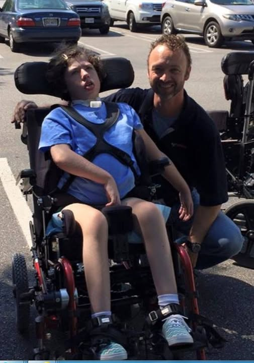 A man kneels and smiles beside a child in a motorized wheelchair in a parking lot, sharing a joyful moment. The scene captures the warmth of Carolines Story, with parked cars in the background. The child wears a blue shirt and shorts; the man wears dark colors.