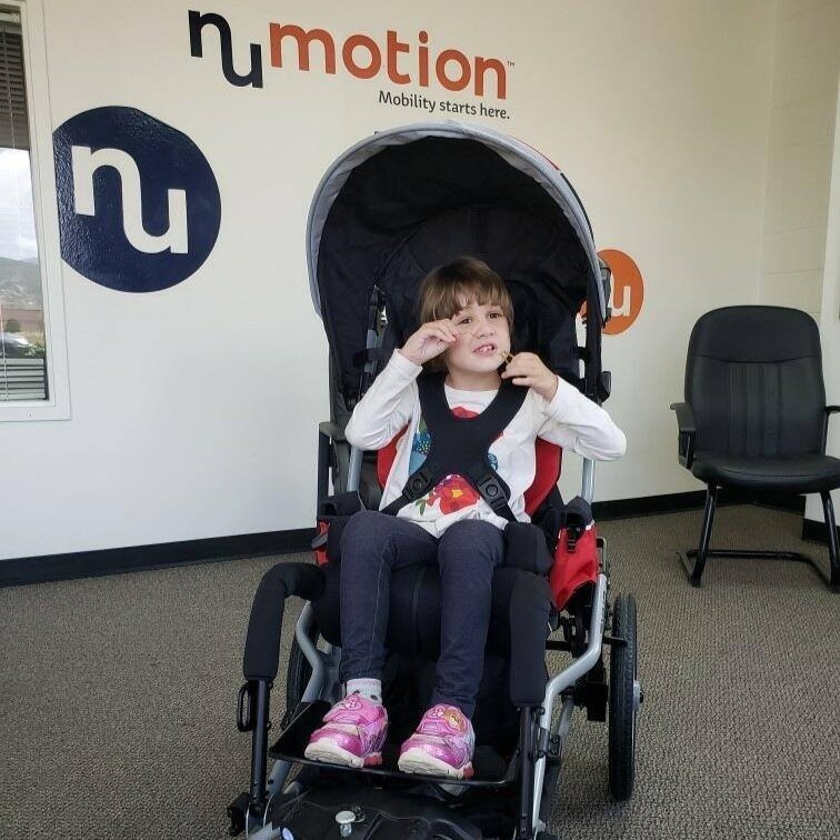 A young girl sits in a specialized wheelchair inside an office with the Numotion logo behind her. She smiles and makes a playful gesture with her hands, capturing a joyful moment from Marleys Story.
