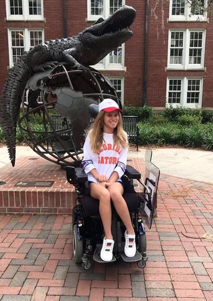 Delainas Story: A young woman in a wheelchair smiles in front of a large gator sculpture and globe at an outdoor campus plaza. She wears a Florida Gators shirt, white hat, and white shoes, with brick buildings and greenery in the background.