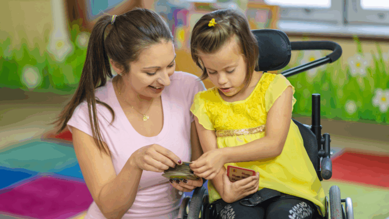 A young woman and a girl using early mobility supports play a card game together in a colorful classroom. Both are smiling and focused on the cards, sharing a joyful and engaging moment while moving to learn.