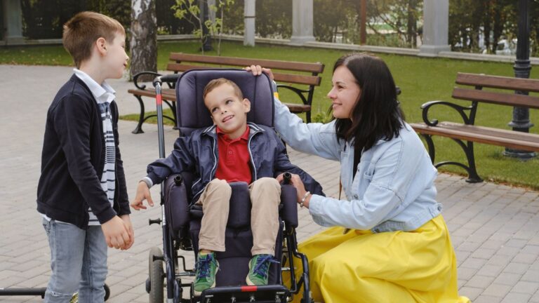 A woman kneels next to a boy seated in a complex rehabilitation stroller, gently touching his head. Another young child stands nearby, smiling at them. They are outdoors in a park setting with benches and greenery.