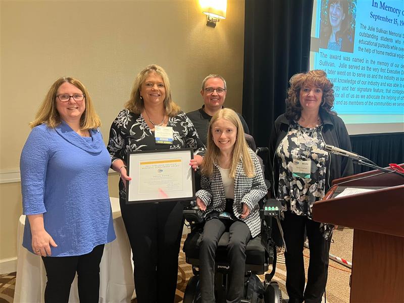 Five people pose together indoors, with one woman seated in a motorized wheelchair holding a framed certificate. A memorial tribute and photo are projected on the screen behind them. Everyone smiles warmly.