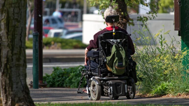 A person with short gray hair uses a motorized wheelchair with a custom configuration on a sidewalk, carrying a green backpack. Trees and greenery surround the path on a sunny day.