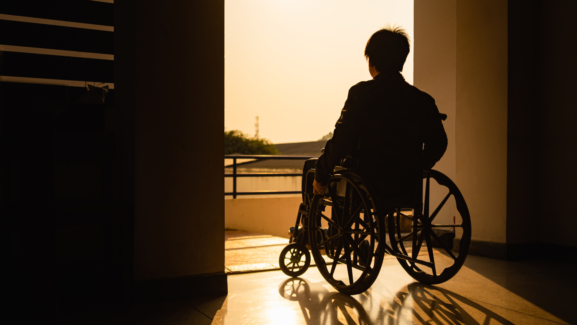 A person using a loaner wheelchair sits in a doorway, gazing out at the sunset. Warm light casts long shadows on the floor, creating a calm and contemplative atmosphere.
