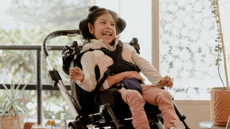 A young girl with dark hair smiles and laughs while sitting in a pediatric powered mobility device indoors, surrounded by plants and natural light from large windows.