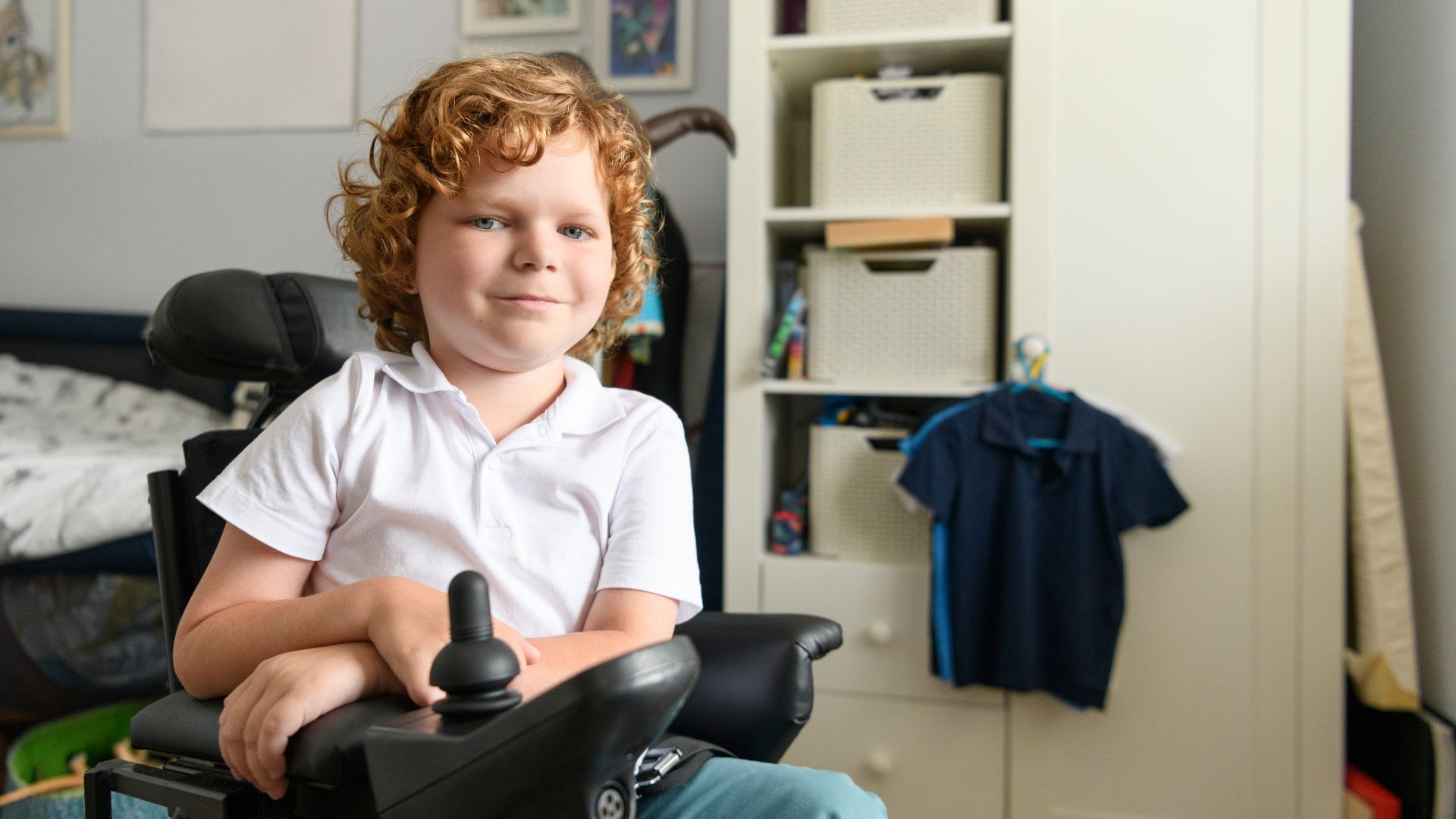 A young child with curly hair sits in a powered mobility device, smiling warmly in a bedroom with a white wardrobe and clothes hanging on hooks in the background—a moment highlighting NuDigest’s focus on parent perception.