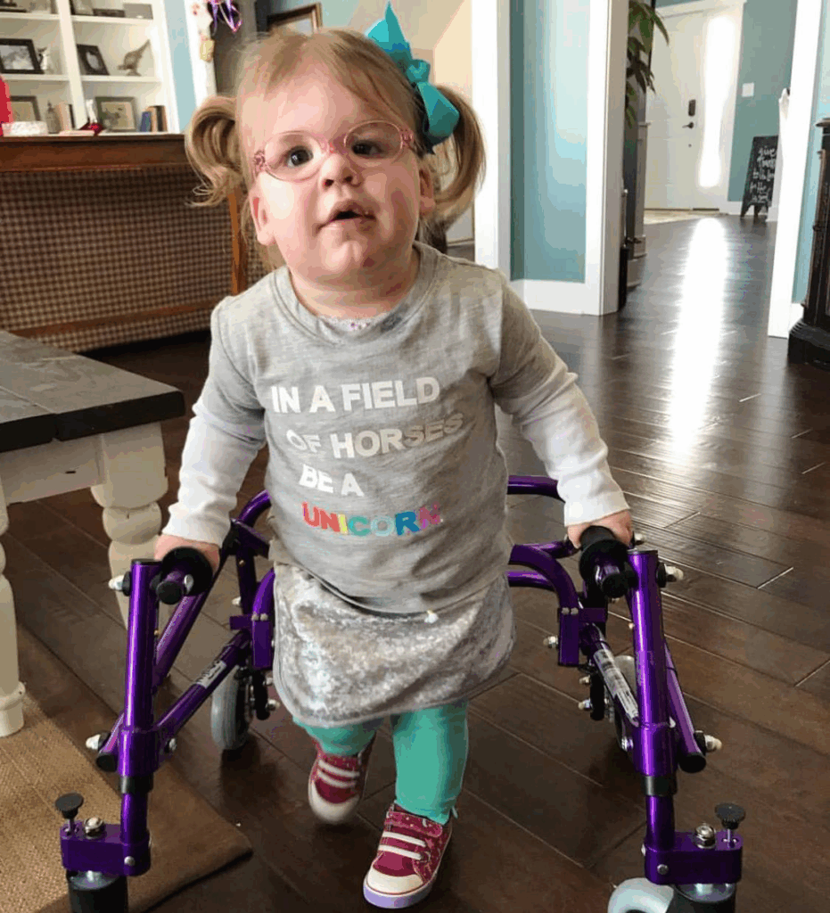 A young girl with glasses and pigtails uses a purple walker in a bright room. Her shirt reads “IN A FIELD OF HORSES BE A UNICORN.” Londyn’s Story shines through as she moves forward, blue bow in her hair, looking up with determination.