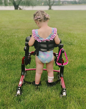 A young child with braided hair, wearing a colorful swimsuit and orthopedic brace, stands on grass using a red walker. Emerys Story unfolds by a serene lake with trees in the background.