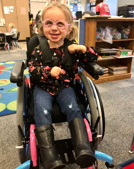 Emerys Story: A young girl with blonde hair, wearing glasses, a black shirt with pink hearts, jeans, and black boots, smiles while sitting in a wheelchair in a classroom setting.