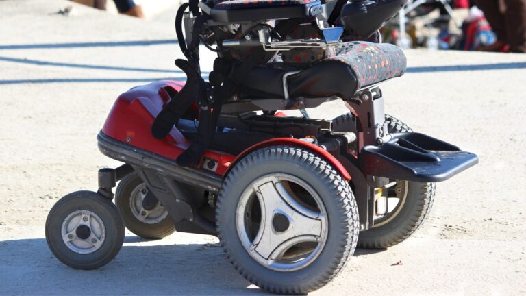 A close-up of a red and black powered wheelchair on a sunny outdoor surface, highlighting large wheels, footrests, and part of the seat, with emphasis on wheelchair safety features.