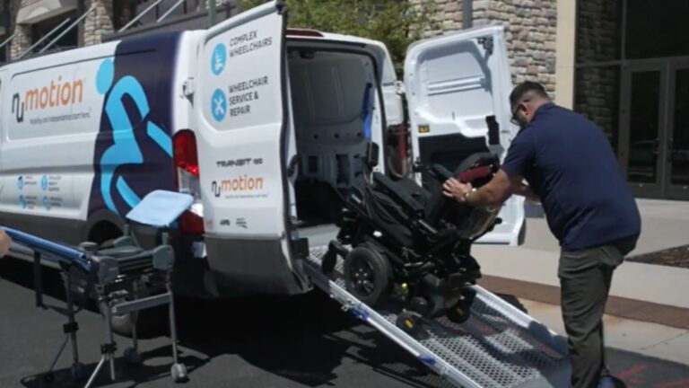 A man pushes a motorized wheelchair up a ramp into a white service van labeled “nMotion,” featuring graphics and text for complex rehab, wheelchair service, and repair. Equipment is visible outside the van.