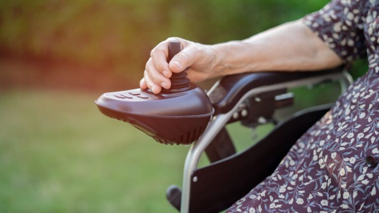 A person’s hand operates the joystick control of an electric wheelchair outdoors. The person, dressed in a patterned dress, uses complex rehab seating for enhanced support. The background is green and blurred.