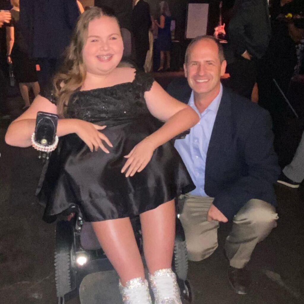 A girl in a black dress sits in a wheelchair, smiling next to a man in a blue jacket and light pants who is crouching beside her. Carlees Story shines through as they enjoy an indoor event with others mingling in the background.