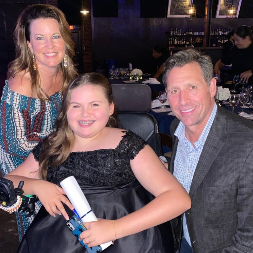 Three people posing and smiling at a restaurant: a woman in a patterned dress, a girl in a fancy black dress holding a rolled paper from Carlees Story, and a man in a plaid suit jacket. Tables with white plates and glasses are in the background.