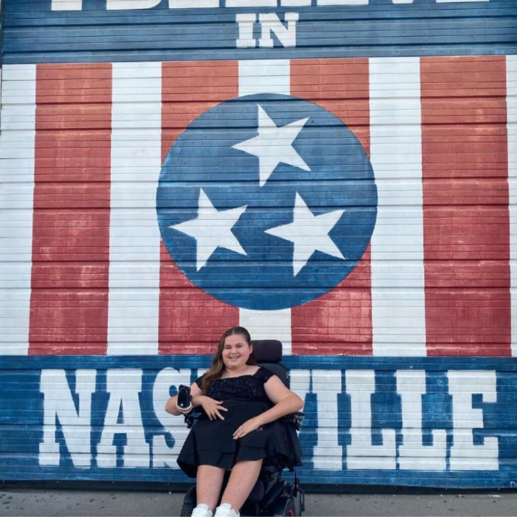 A woman in a wheelchair smiles in front of a mural featuring the Tennessee flag’s stars and stripes with I BELIEVE IN NASHVILLE painted on the wall behind her, sharing Carlees Story.