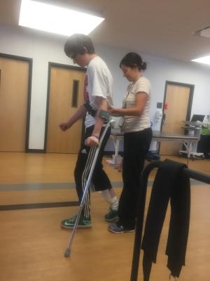 A young person using crutches is assisted by an adult during physical therapy in a room with wooden floors and closed doors, illustrating Ryans Story.