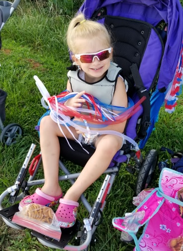 A young girl with blonde hair in a ponytail sits in a purple stroller, smiling. She wears pink sunglasses, a light top, and pink Crocs, holding red, white, and blue ribbon wands on her lap—this is part of Haydens Story.