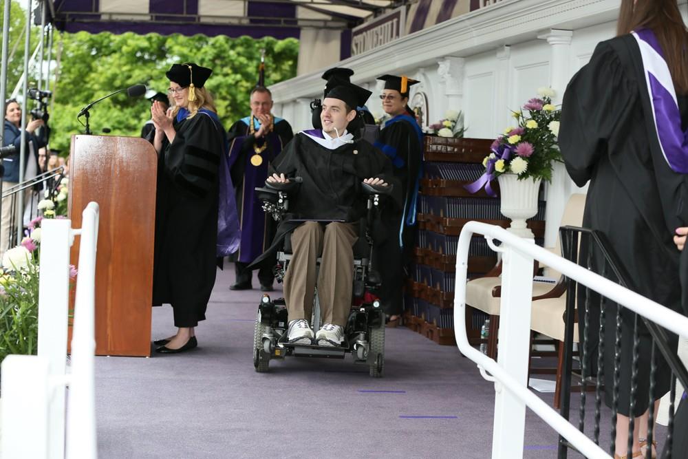 A graduate in a wheelchair, wearing a cap and gown, smiles on stage during an outdoor graduation ceremony. Matts Story shines as faculty in academic regalia stand nearby, with flowers decorating the stage.