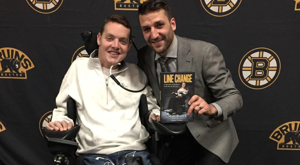 Two men pose in front of a Boston Bruins backdrop; one, seated and holding a book titled “Line Change,” shares Matts Story, while the other, in a suit, smiles and points at the book.