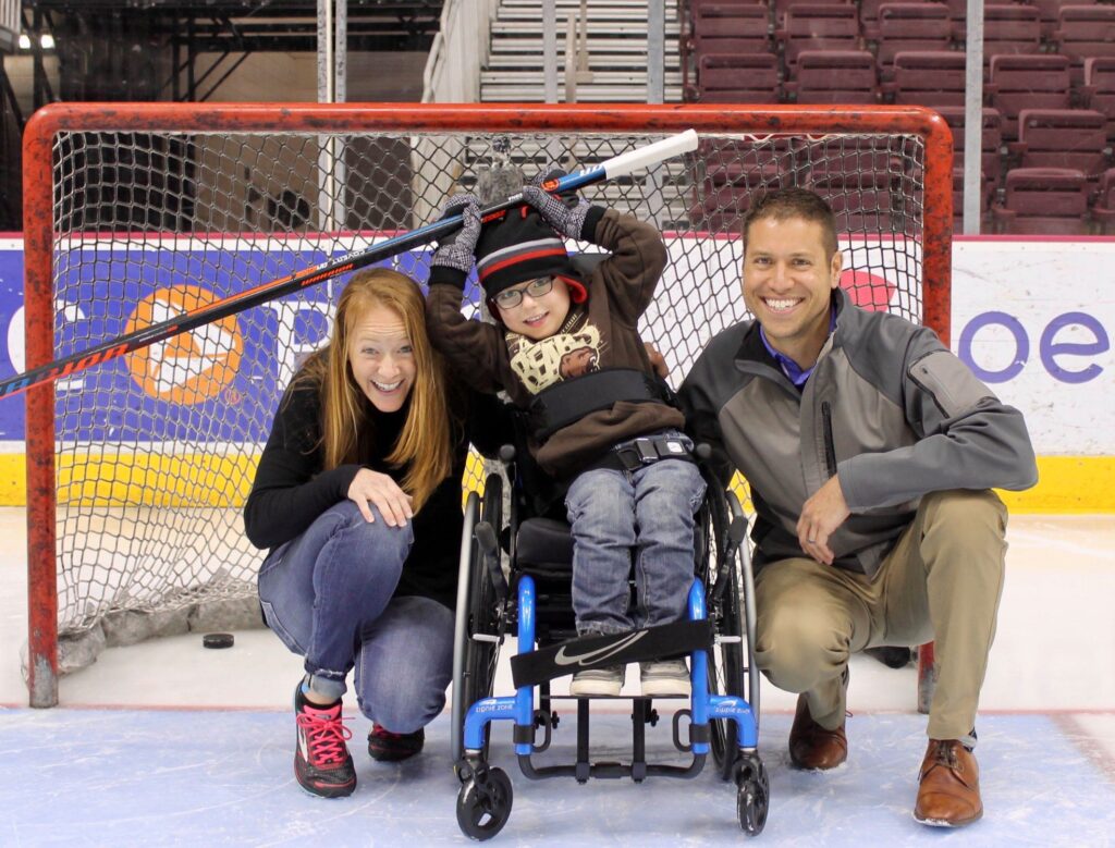 A smiling woman and man kneel beside a boy in a wheelchair holding a hockey stick, all posing happily in front of a hockey net on an ice rink—capturing the spirit of Parkers Story.