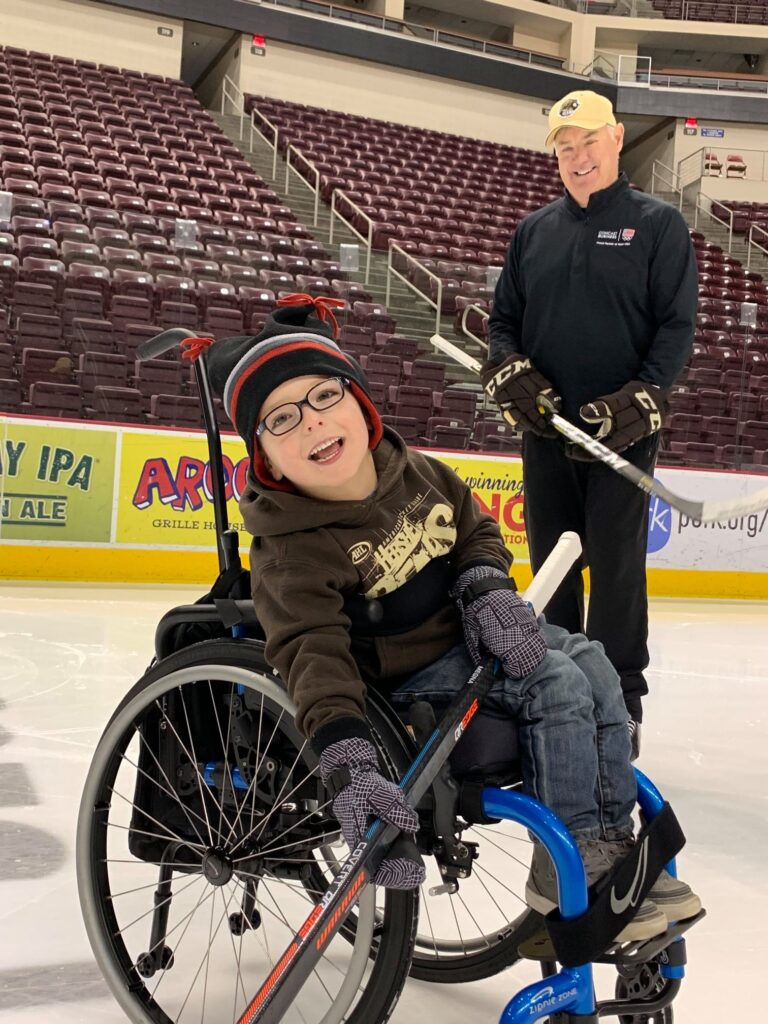 A smiling child in a wheelchair holds a hockey stick on an ice rink, with an adult in hockey gear standing behind, also smiling. The empty arena seats set the scene for Parkers Story.