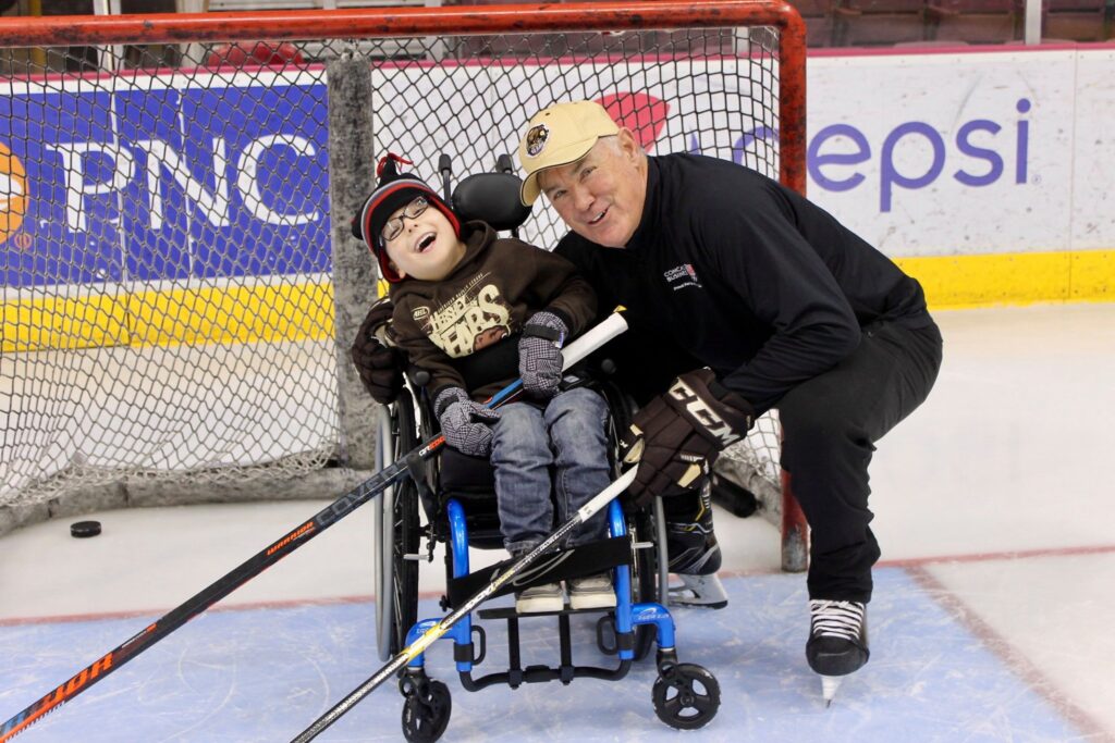 A smiling man in hockey gear kneels beside a young boy in a wheelchair on an ice rink. Both hold hockey sticks and pose in front of the goal net, enjoying the moment together—an inspiring scene from Parkers Story.