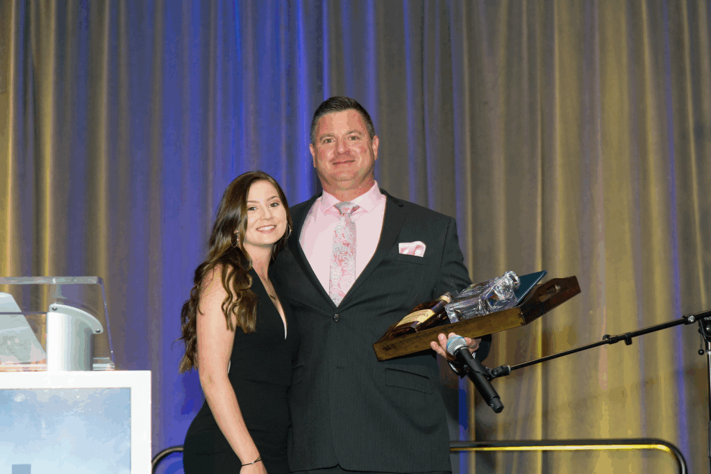 A man in a suit holding a wooden award plaque stands next to a woman in a black dress. They are smiling and posing for a photo at an event with gold and blue curtains, celebrating Todds Story.