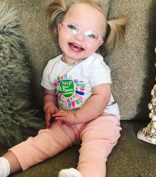A smiling young child with glasses and pigtails sits on a couch, wearing a T-shirt with colorful designs and light pink pants. The child looks up, appearing joyful and happy—a delightful moment from Hatties Story.