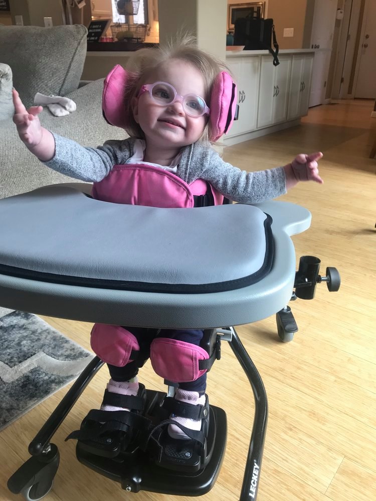 A smiling toddler wearing glasses and a padded pink support vest sits in a supportive chair with leg braces and straps, arms outstretched on a wooden floor. Part of Hatties Story.