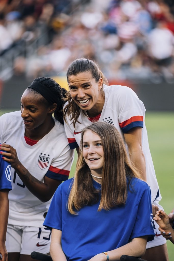 Three women at a soccer field: two wear white USA soccer jerseys and smile, while a young woman in front, wearing a blue shirt, also smiles—capturing a joyful moment from McKennas Story with a blurred crowd in the stadium background.