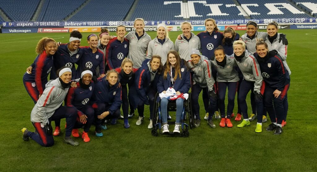 A women’s soccer team poses on a field with a smiling young fan at the center, celebrating McKennas Story. The players and coaches, dressed in team gear, gather closely to show unity and support.