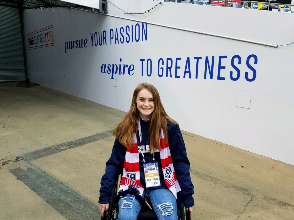 A smiling girl wearing a soccer scarf and credentials sits in a wheelchair in front of a wall with the words pursue YOUR PASSION aspire TO GREATNESS at a sports venue, celebrating McKennas Story.