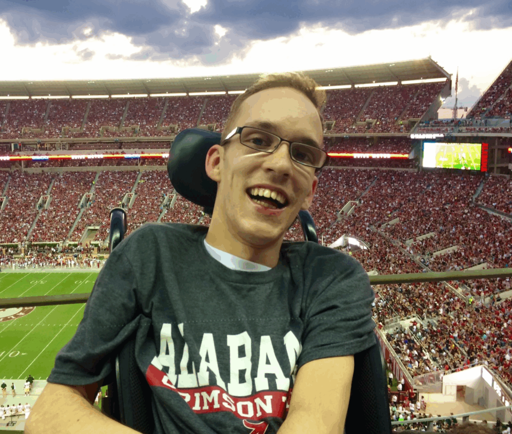 Seths Story: A young man wearing glasses and an Alabama Crimson Tide shirt smiles while sitting in a stadium packed with fans during a football game, with the field and stands visible in the background.