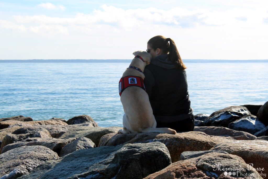 Katelynne sits on a rocky shore beside a yellow lab in a red service vest, both facing a calm, expansive body of water under a bright sky, sharing a peaceful moment that hints at their unique story.