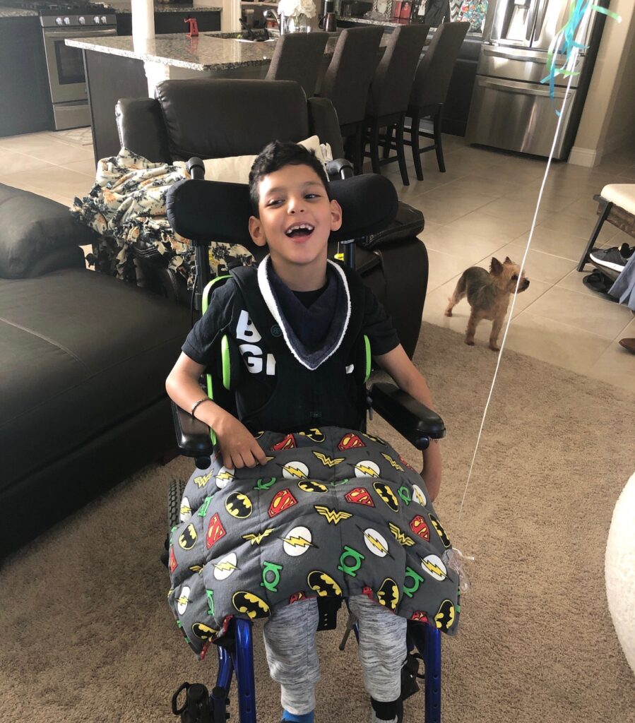 A smiling boy sits in a wheelchair with a superhero-themed blanket on his lap, part of Jojos Story. In a cozy living room, a small brown dog stands nearby, with the kitchen and dining area visible in the background.