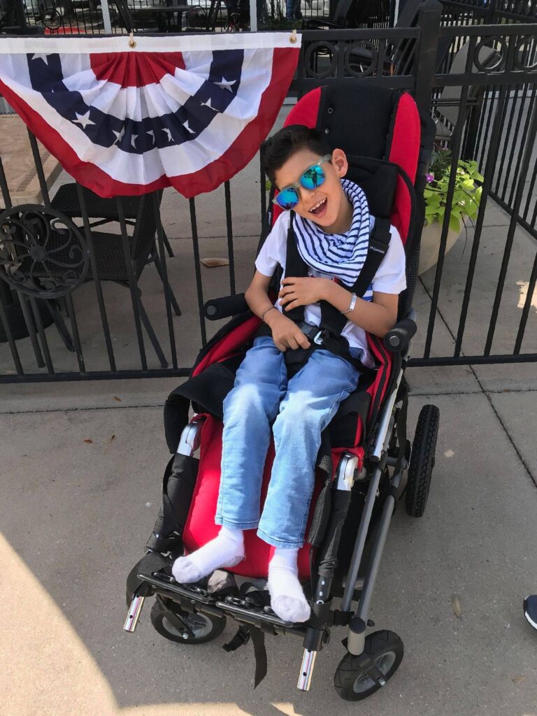 A smiling boy sits in a red wheelchair, wearing sunglasses, a striped scarf, jeans, and a white shirt. Behind him is a black fence adorned with a red, white, and blue patriotic banner—a joyful moment from Jojos Story.