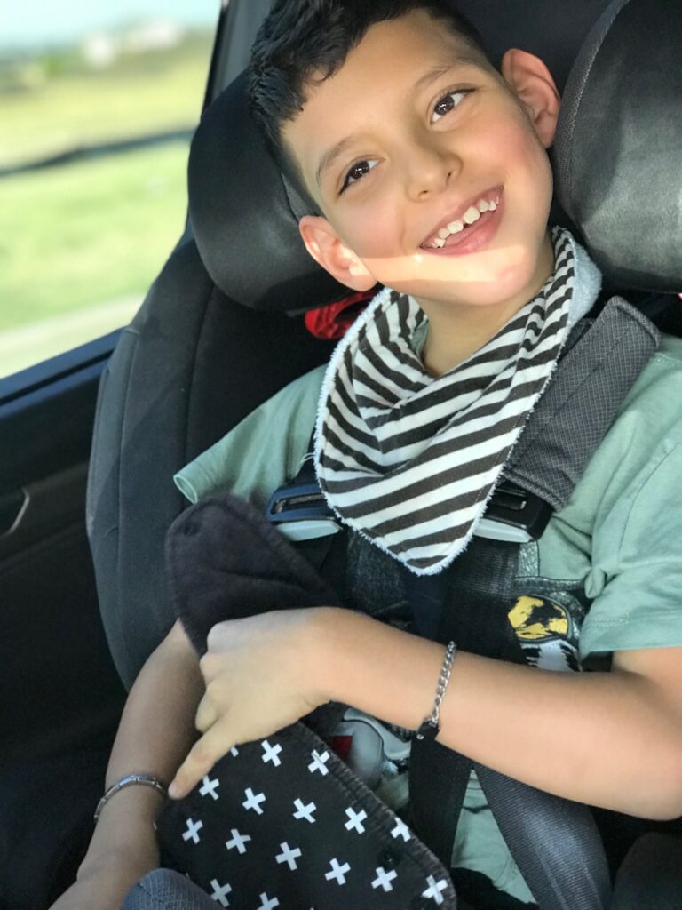 A young boy with short hair smiles while sitting in a car seat. Wearing a green shirt and striped bib, he holds a black pillow with white crosses. Sunlight brightens his face as he looks out the window, adding joy to Jojos Story.