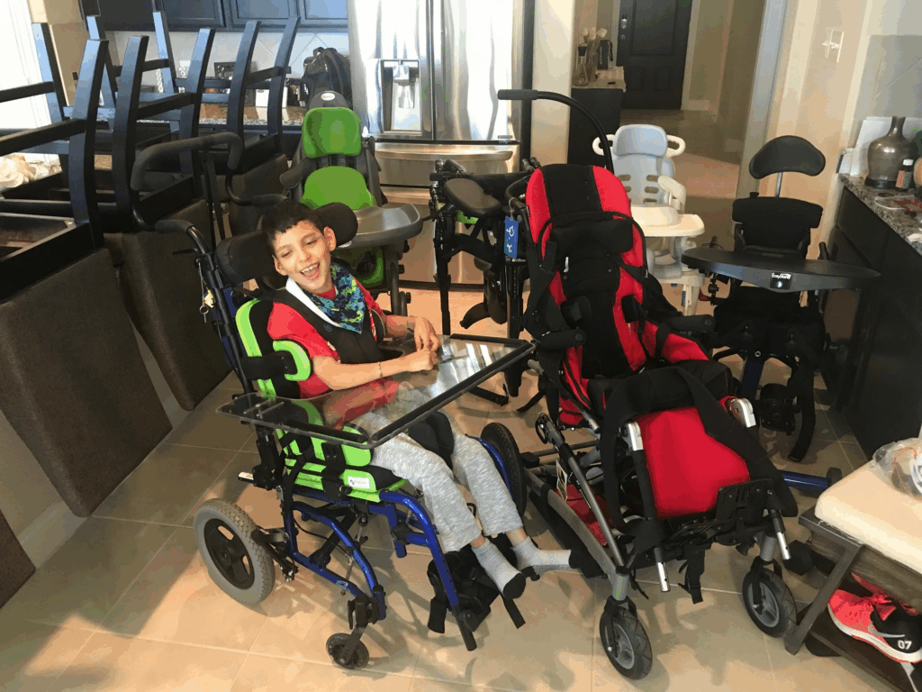 A smiling young boy sits in a green and blue wheelchair with a clear tray in a kitchen, surrounded by several other colorful wheelchairs and high chairs. Jojos Story comes to life amid the vibrant atmosphere and stacked tables in the background.