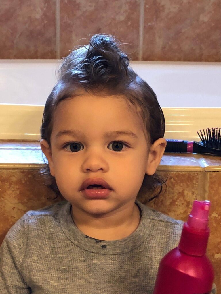 A young child with curly hair styled into a small mohawk gazes at the camera, holding a pink spray bottle. Zanders Story unfolds as a hairbrush rests in the background on a tiled bathroom surface.