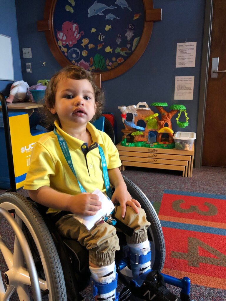 Zander, a young child in a wheelchair, wearing a yellow shirt and medical braces on their legs, sits on a colorful rug with large numbers in a playroom decorated with sea-themed wall art and toys—ready for the next story.